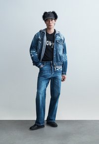 Νεαρός άνδρας με φθαρμένο τζιν μπουφάν και τζιν παντελόνι, μαύρη μπλούζα με σχέδιο, καπέλο με γούνα και μαύρα αθλητικά παπούτσια, στέκεται μπροστά σε απλό φόντο.