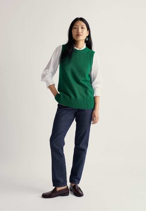 Grüner gestrickter ärmelloser Pullover über einer weißen Bluse, kombiniert mit dunkelblauen Jeans und braunen Schuhen. Einfache strukturierte Musterung; lässige Passform.