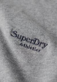 Grijze stof met een zachte textuur heeft donkerblauwe geborduurde tekst die prominent "SuperDry Athletics" op het oppervlak toont.