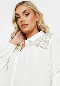 Camicia bianca con bottoni a maniche lunghe plissettate, con dettagli in pizzo sulle spalle e una vestibilità comoda. Tessuto morbido e leggero.
