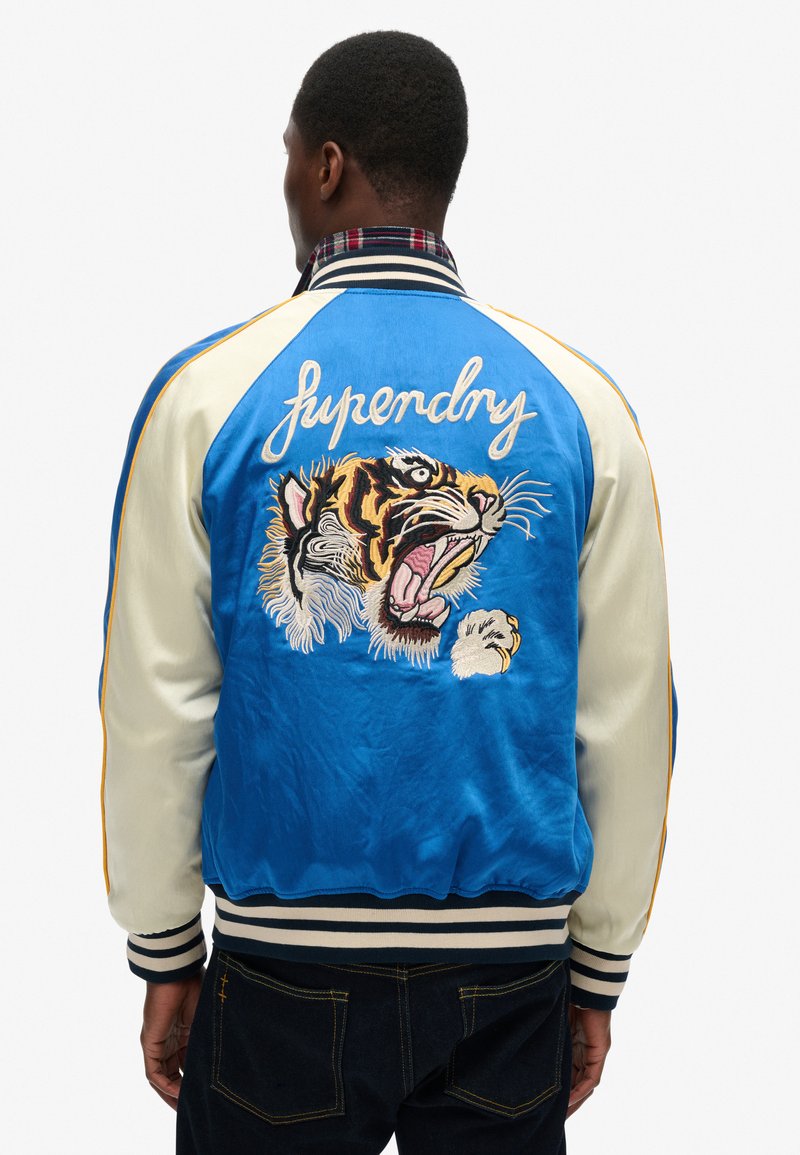 Veste bomber en satin bleu avec des manches crème, présentant un grand tigre brodé et le texte "Superdry" au dos. Poignets et ourlet côtelés.