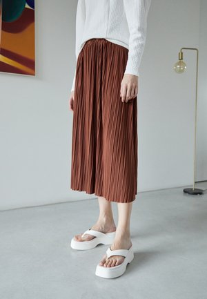 Woman wearing a white sweater, brown pleated midi skirt, and white platform sandals standing indoors near a floor lamp and abstract wall art.