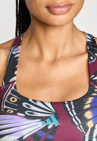 Top deportivo con estampado de mariposas, en colores púrpura, negro y burdeos. Incluye un pequeño logo azul en la parte inferior derecha. Textura suave.
