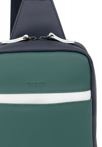 Borsa verde con texture e dettagli blu navy, presenta una striscia orizzontale bianca, doppi cursori per la cerniera e logo impresso "HEXAGONA."