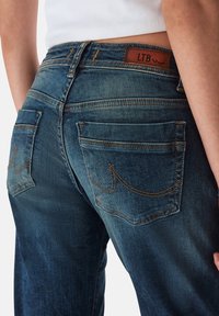Jeans de denim azul oscuro con un acabado desgastado, que cuentan con dos bolsillos traseros, detalles de costura y una etiqueta de marca de cuero marrón.