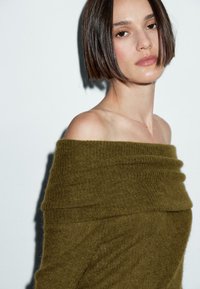 Schulterfreier Pullover in Olivgrün, aus weichem Strickmaterial mit gerippten Details. Glatte Textur und lässiges, drapiertes Design.