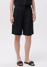 Zwarte linnen shorts met een ontspannen pasvorm, voorzien van voorplooien, zijzakken en een soepele textuur. Gekocht met platte zwarte sandalen.