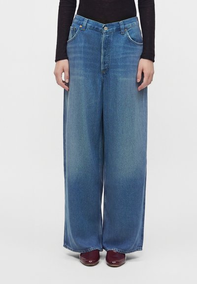 Personne portant un jean bleu taille haute à jambes larges, un haut noir à manches longues et des chaussures bordeaux, debout devant un fond uni.