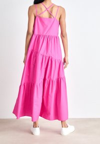 Maxi sukienka w jasnoróżowym kolorze, wykonana z gładkiego materiału. Ma warstwowy krój, cienkie ramiączka z krzyżującym się tyłem oraz białe trampki.