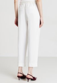 Donna che indossa pantaloni bianchi tagliati con tasche posteriori e décolleté borgogna, in piedi contro uno sfondo chiaro e semplice.