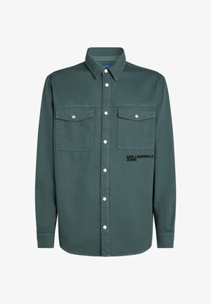 Tummanvihreä nappipaita, joka on valmistettu kudotusta kankaasta ja jossa on kaksi rintataskua sekä hopeiset napit. Vasemmalla puolella on brodeerattu logo.