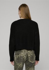 Schwarzer, gerippter Pullover mit kurzem Schnitt. Kombiniert mit olivgrünen Jeans, die einen Leopardenprint und kontrastierende Nähte an den Taschen aufweisen.