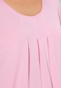Světle růžová blůza s kulatým výstřihem a vertikálními sklady. Tkanina vypadá měkce a lehce, čímž podtrhuje její ležérní styl.