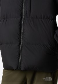 Fekete, teljes cipzárral ellátott puffertdzseki, steppelt mintázattal. Mellkasi zsebbel és bal csípőn logóval rendelkezik. Az anyag fényes és szigetelt hatású.