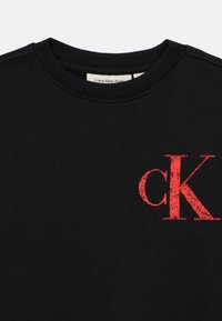 Svart sweatshirt av bomull, med ribbad halsringning och en stor, sliten röd "CK"-logotyp på framsidan.