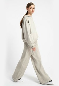 Młoda kobieta idąca, ubrana w oversize'owy beżowy hoodie i pasujące szerokie spodnie, w białych sneakersach, na jednolitym białym tle.