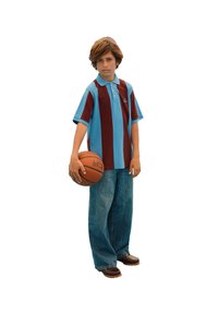 Deček, oblečen v modro in maroon črtasto polo majico in široke kavbojke, drži košarko v eni roki, stoji na beli podlagi.