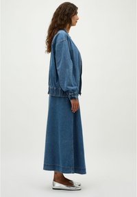 Chaqueta de denim con puños elásticos, combinada con una larga falda de denim. Zapatos planos grises. Todos los artículos en diferentes tonos de azul. Textura suave.