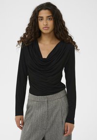 Sieviete ar gariem, cirtainiem matiem, tērpta melnā drapētā garās piedurknes topā un pelēkām rakstainām augsto jostasvietu biksēs uz neitrāla fona.