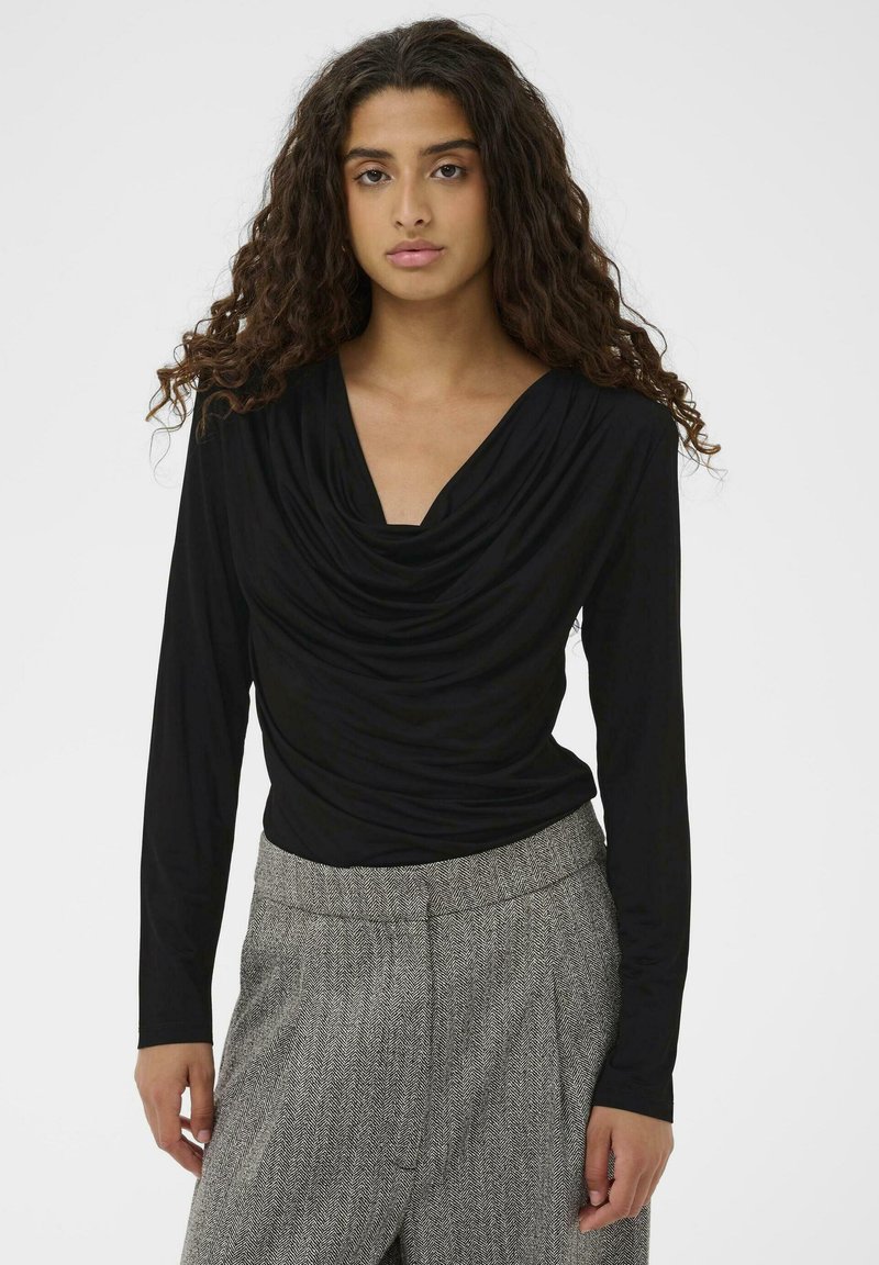 Sieviete ar gariem, cirtainiem matiem, tērpta melnā drapētā garās piedurknes topā un pelēkām rakstainām augsto jostasvietu biksēs uz neitrāla fona.