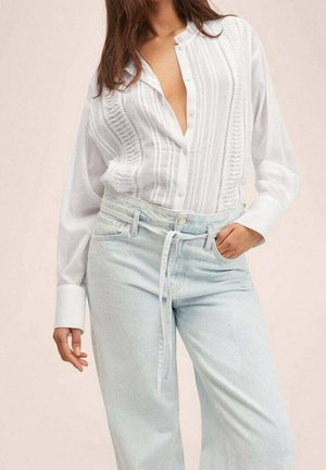 Frau in weißer Bluse mit Knöpfen und strukturierten Streifen, in hellblaue, hoch taillierte Jeans mit Stoffgürtel gesteckt, Hand in der Hosentasche.