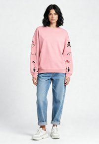 Felpa rosa con maniche lunghe e design ricamato nero. Abbinata a jeans azzurri a risvolto e sneakers bianche.