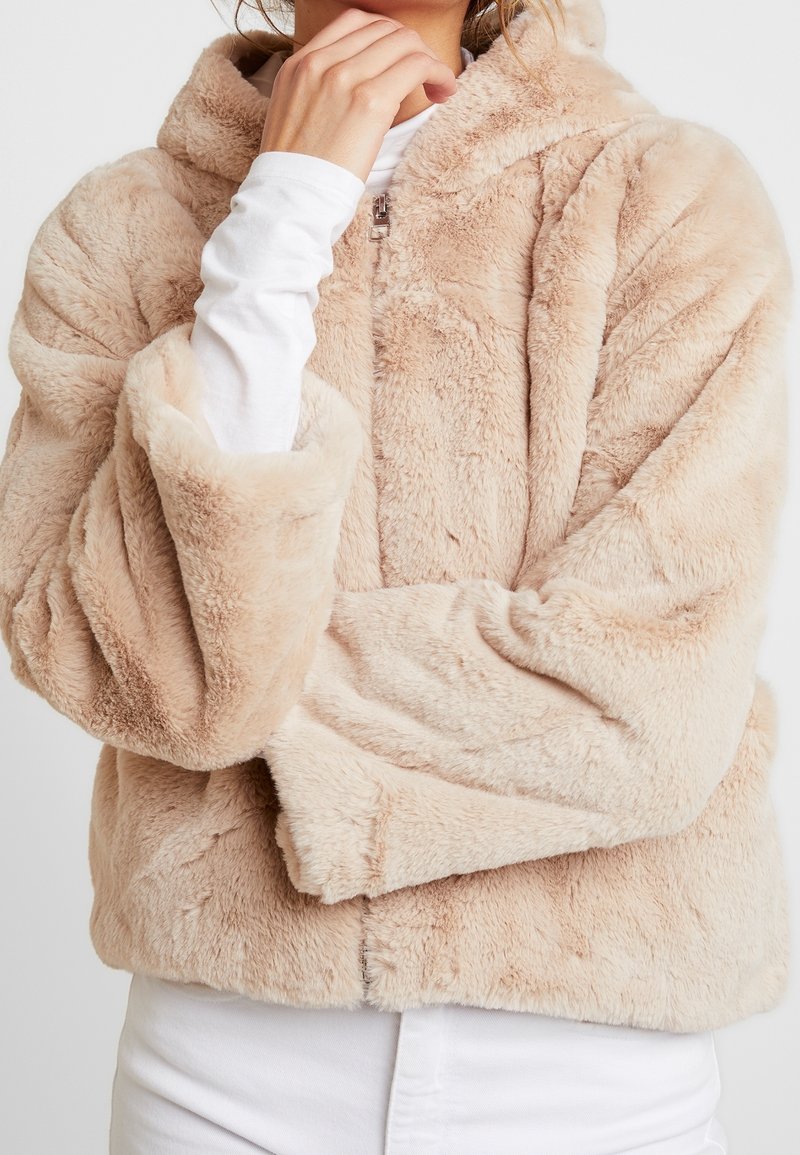 Beżowa bluza z kapturem z sztucznego futra z zamkiem błyskawicznym, obszerne rękawy, pluszowa faktura i przytulny kaptur, noszona na białej bluzce z długim rękawem.