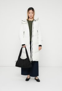 Hvid pufferjakke med hætte, med en frontlynlås og sidelommer. Båret over en grøn sweater, parret med brede mørke jeans og en sort taske.