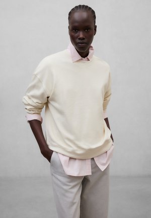 Sort person med fletninger iført cremefarvet oversized sweatshirt over lys pink skjorte og beige bukser, stående med hænderne i lommerne mod en ensfarvet baggrund.