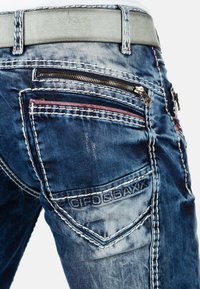 Jeans in denim con un lavaggio blu sfumato, caratterizzati da numerosi tasche, cuciture a contrasto e dettagli testurizzati. Tasca con zip e cucitura decorativa.