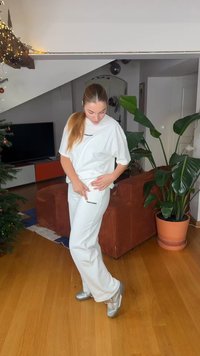 Άσπρο φαρδύ t-shirt με μαύρο κείμενο, συνδυασμένο με αντίστοιχο άσπρο παντελόνι. Ασημένια παπούτσια με γυαλιστερό φινίρισμα. Εσωτερικός χώρος με φυτά.