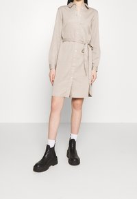 Lyhythihainen, pitkähihainen beige verkkovaatetus, jonka edessä on napitus ja vyötäröllä solmittava vyö. Yhdistetty mustiin paksupohjaisiin nilkkureihin ja valkoisiin sukkiin.