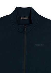 Veste zippée marine en tissu lisse avec col montant. Présente un logo embossé discret et une fermeture éclair contrastante.