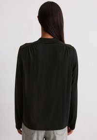 Haut noir à manches longues avec une texture lisse, présentant un détail froncé subtil aux épaules et un col rond, associé à un pantalon gris clair.