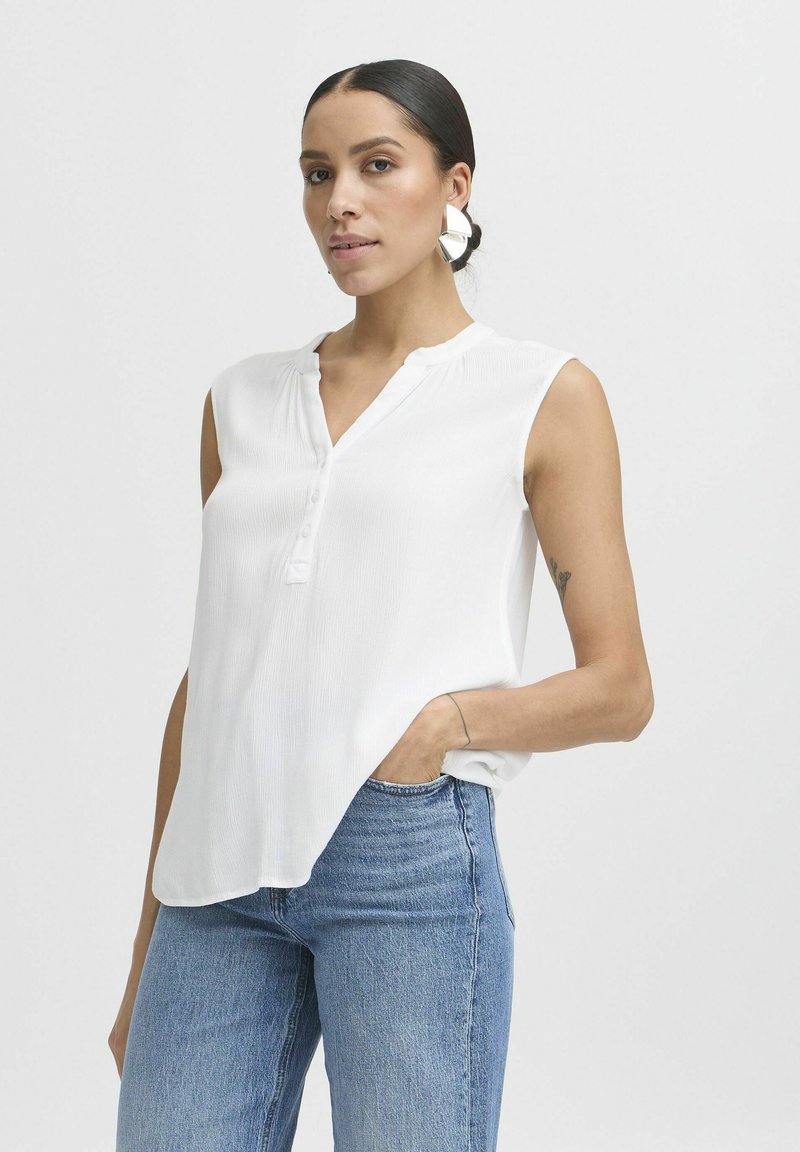 Weißes ärmelloses Blusenshirt mit Henley-Ausschnitt, strukturierter Stoff und drei Knöpfen vorne. Kombiniert mit blauen Jeans.