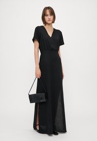 Melnas maksi kleitas ar īsām piedurknēm, apvērsta dizaina un sānu šķeltnēm. Modelis tur melnu crossbody somu ar ķēdes siksnu. Gluda tekstūra.
