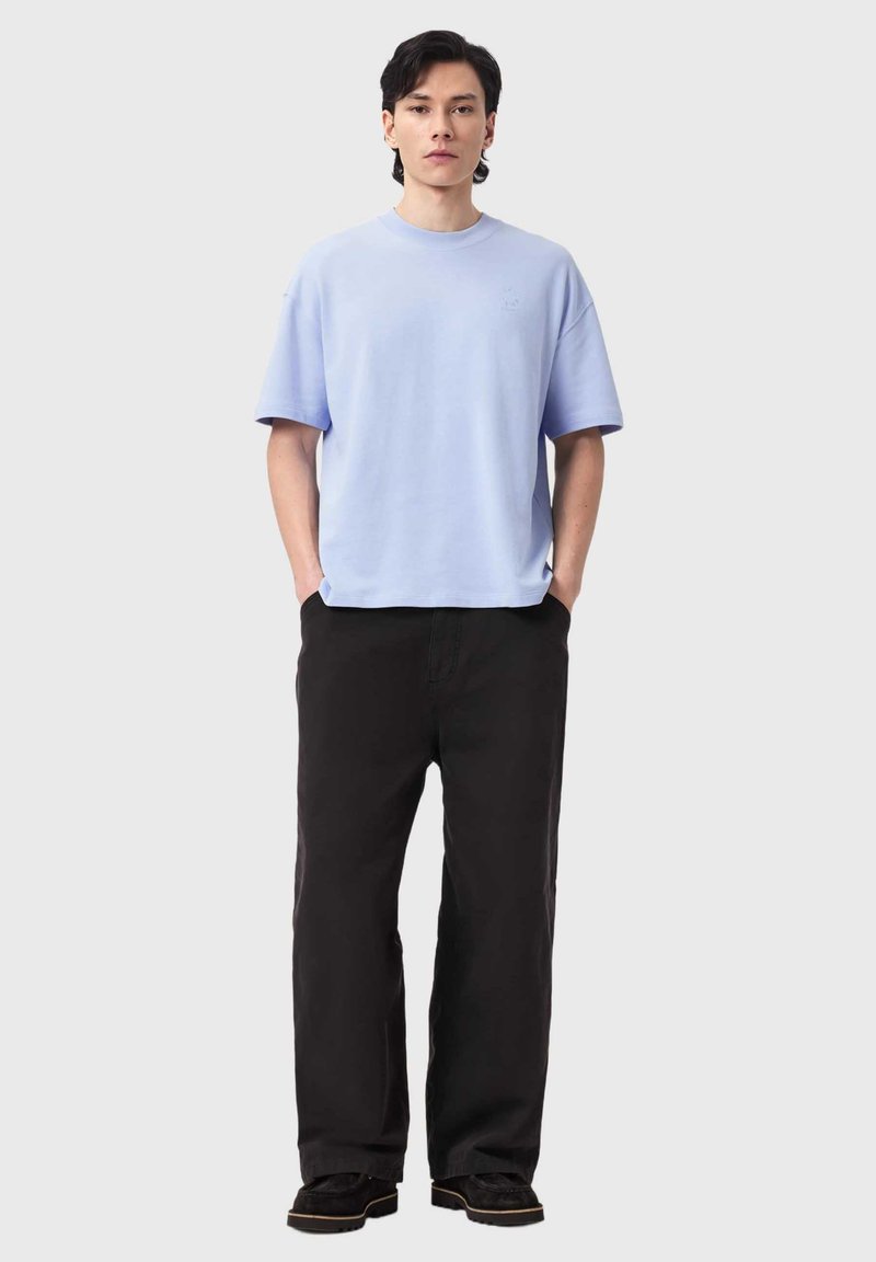 Junger Mann steht mit den Händen in den Taschen, trägt ein hellblaues, übergroßes T-Shirt, schwarze weit geschnittene Hosen und schwarze Schuhe vor einem schlichten Hintergrund.