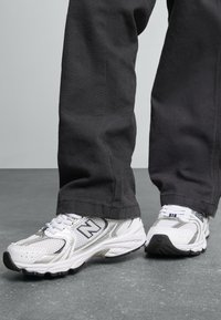Baskets de sport blanches avec des accents argentés et des panneaux en maille, dotées d'une semelle extérieure noire, associées à un pantalon sombre et ample.