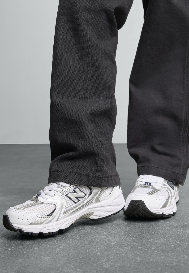Baskets de sport blanches avec des accents argentés et des panneaux en maille, dotées d'une semelle extérieure noire, associées à un pantalon sombre et ample.
