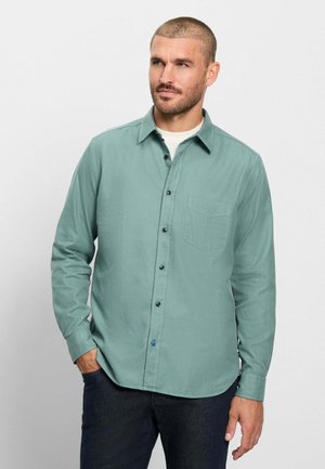 Mann mit kurzen Haaren, der ein grünes Hemd mit Knöpfen über einem weißen T-Shirt trägt, steht mit einer Hand in der Tasche vor einem schlichten hellen Hintergrund.