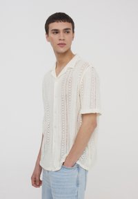 Camicetta leggera di colore crema a maniche corte con trama a maglia, dotata di chiusura a bottoni e vestibilità rilassata.