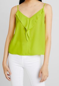 Top verde, ligero y sin mangas con escote en V y detalle asimétrico de volante en la parte delantera, combinado con pantalones blancos.