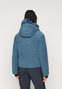 Modrá kordurová bunda s podšívkou kapuce, disponující vroubkovanou strukturou a jednoduchým designem. Zadní pohled ukazuje detaily šití.