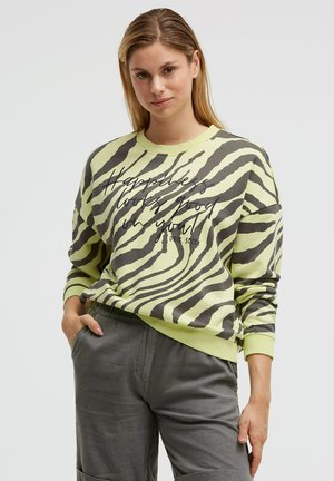 Woman with light brown hair wears yellow-green zebra-striped sweatshirt with "Happiness looks good on you" text and gray pants, posing.