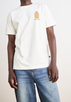 Mand iført en hvid t-shirt med en lille tønde og "new balance" logo på brystet, kombineret med blå falmede jeans.