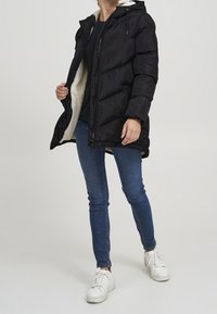 Svart puffjacka med huva, med en mjuk vit foder, dragkedjestängning och sidofickor, i kombination med blå jeans och vita sneakers.