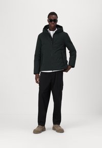 Black hooded jacket with a front zipper, worn over a white shirt. Paired with black cargo pants and beige shoes. Sunglasses worn.