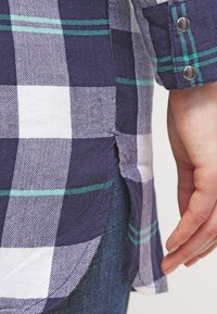 Navy and white checked button-up shirt with herringbone texture, featuring green accent stripes and a side slit. Hand resting near the hem.