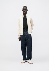 Lys beige bomberjakke over en svart t-skjorte, kombinert med løstsittende blå jeans og hvite joggesko med brune såler, stående på en ensfarget bakgrunn.