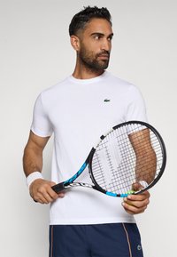 T-shirt bianca a girocollo con un piccolo logo verde, indossata da un uomo che tiene una racchetta da tennis e una palla, caratterizzata da maniche corte e una superficie liscia.